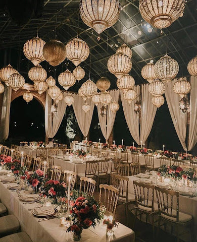 Beautifully set dining tables and chairs inside a venue at night with lanterns providing overhead lighting.