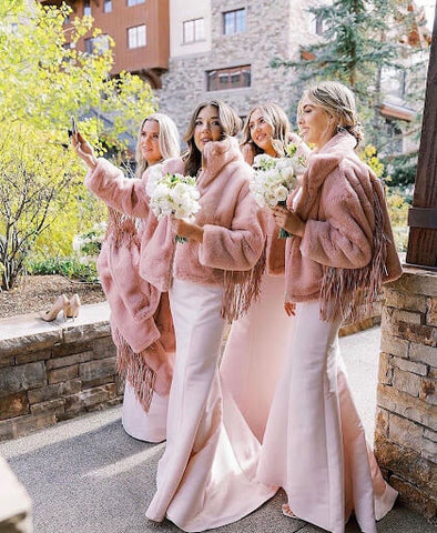 Four fabulous bridesmaids in pale pink dresses with luxuriously fuzzy wraps and white flower bouquets taking a selfie.
