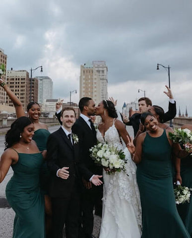 Bride and groom kissing while their wedding party stands celebrating around them. City in the background.