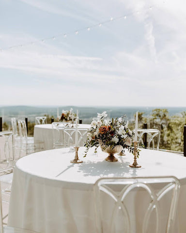 Round tables with white tablecloths and clear plastic chairs at an upscale outdoor patio venue under blue skies.