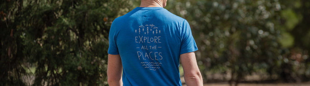 Model wearing ioMerino's Men's Universal Tee in Dazzling Blue with Explore all the Places print, standing in front of shrubbery.