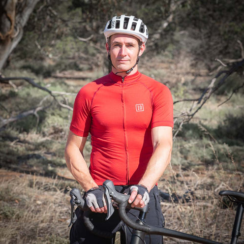 Model wearing ioMerino's Men's Cycling Jersey in Chilli and a helmet, standing on a trail with a bike.
