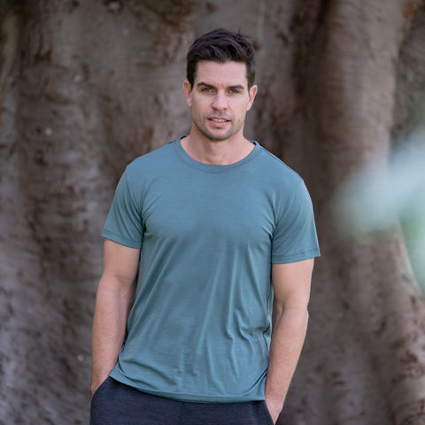 Model wearing ioMerino's Men's Universal Tee in Silver Pine, standing with hands in pants pockets in front of a tree