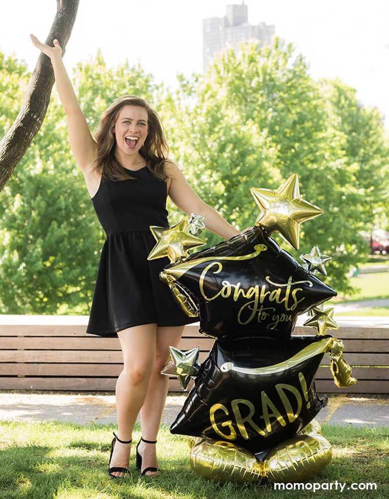 A high school or college graduation standing next to Momo Party's 51" Airloonz Congrats To You Grad Foil Mylar Balloon by Anagram Balloons. Her hand is up in the air celebrating her big achievement on the graduation day.