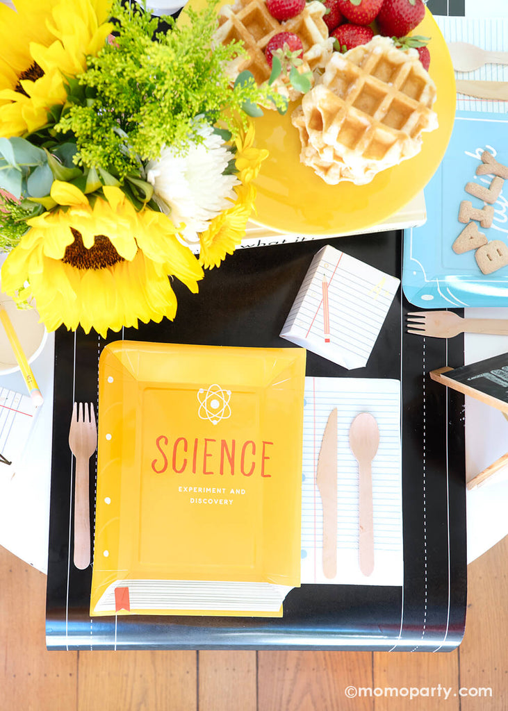 An overhead shot of a back to school party tablescape featuring multiple school themed party goods from Momo Party, including text book shaped plate in different yellow, the notebook napkin and a treat box, all on the chalkboard table runner. With stacked waffles on a cake stand, fruits, alphabet cookies, and a sunflower floral arrangement on the table, this makes a perfect idea for your child's back to school party or first day of school celebration!