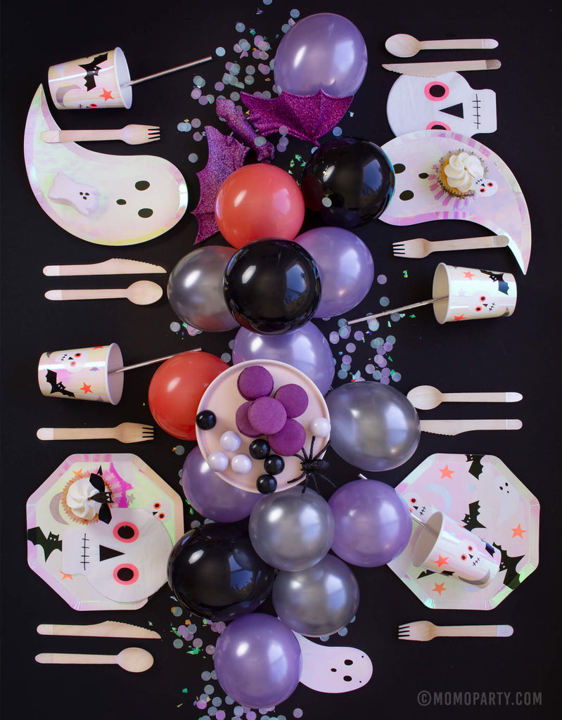 Momo Party Halloween Party table set up with halloween icon plates, iridescent ghost plates, skull napkins, and halloween icon cups, wooden utensils on the tables,  purple coral latex balloons as centerpiece 
