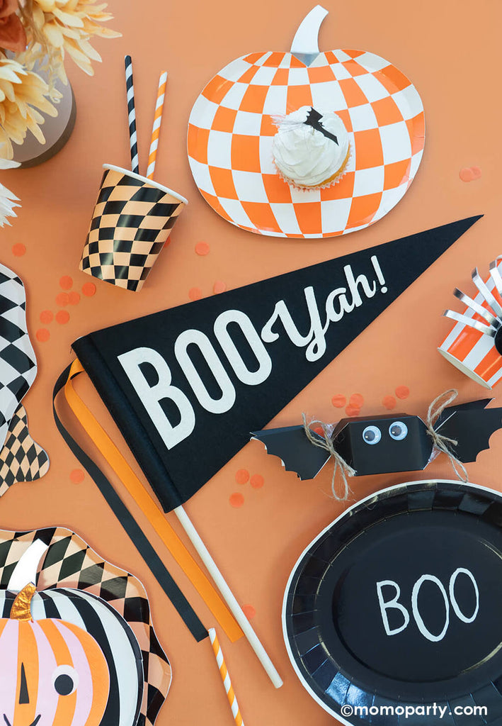 A Halloween party table featuring Momo Party's felt Boo-yah party pennant, around it there are swirling checkered plates, cups, napkins, pumpkin shaped plates with googly eyes, bat shaped jelly beans, black tombstone shaped napkins and boo shaped plates, all makes a spooky yet fun decoration ideas for a Halloween bash this season.
