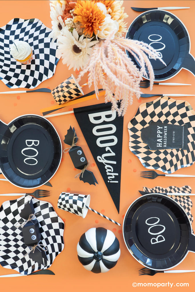 A table filled with Momo Party Classic Orange and Black Halloween Party Decorations including swirling checkered plates, cups, napkins, Boo Yah party pennant, bat shaped jelly beans, black tombstone shaped napkins and boo shaped plates, all makes a spooky yet fun decoration ideas for a Halloween bash this season.