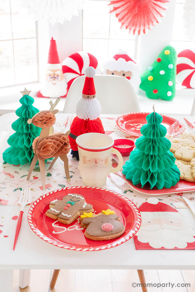 A festive Christmas cookie decorating party table featuring Momo Party's Cookies for Santa red round plates, Santa face party cups, Santa shaped napkins and red and white cutlery set. In the back, there are Christmas character honeycomb featuring Santa, Christmas trees and reindeers.