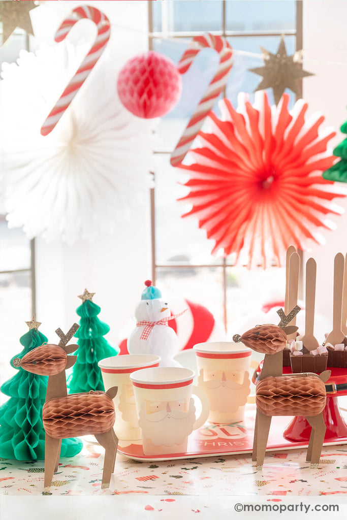 A festive Christmas party table featuring a hot cocoa bar with Momo Party's Santa hand cups with handles, hot chocolate stirrer spoons and Christmas honeycomb decorations of Santa, snowman, reindeers and Christmas trees. In the back there's a jumbo Christmas honeycomb garland with candy canes, peppermint paper fans and Christmas trees.