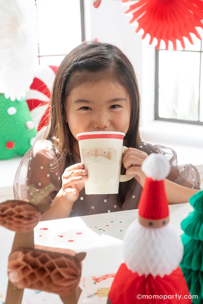 A girl in her Christmas outfit holding a Santa face shaped party cup with hot cocoa in a Christmas cookie decorating party decorated with Momo Party's honeycomb decorations featuring Santa, reindeers and Christmas trees.
