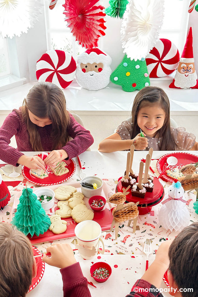 Four kids at a Christmas cookie decorating party which is decorated with festive holiday decorations like honeycomb, paper fans and Christmas themed tableware from Momo Party. Kids are dress in their holiday outfit using pippin tube to decorate their holiday cookies - a great festive activity or playdate idea for children during the holiday season.