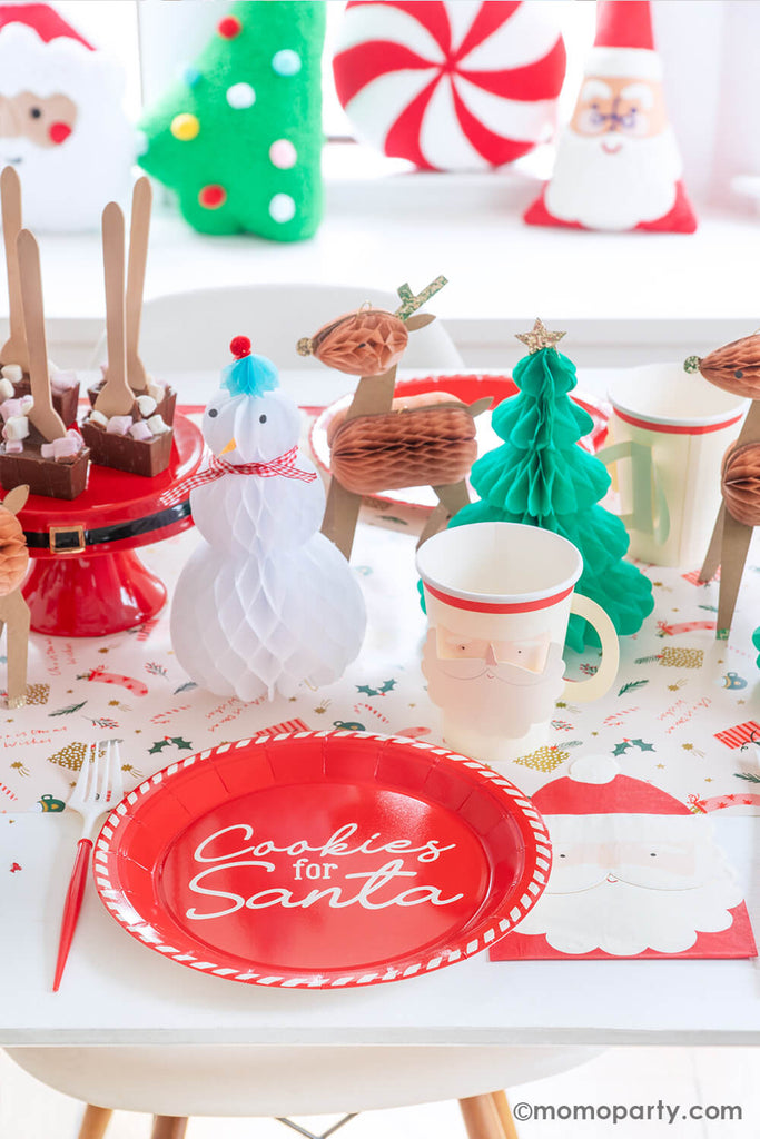 A festive Christmas cookie decorating party table featuring Momo Party's Cookies for Santa red round plates, Santa face party cups, Santa shaped napkins and red and white cutlery set. In the back, there are Christmas character honeycomb featuring Santa, Christmas trees and reindeers.