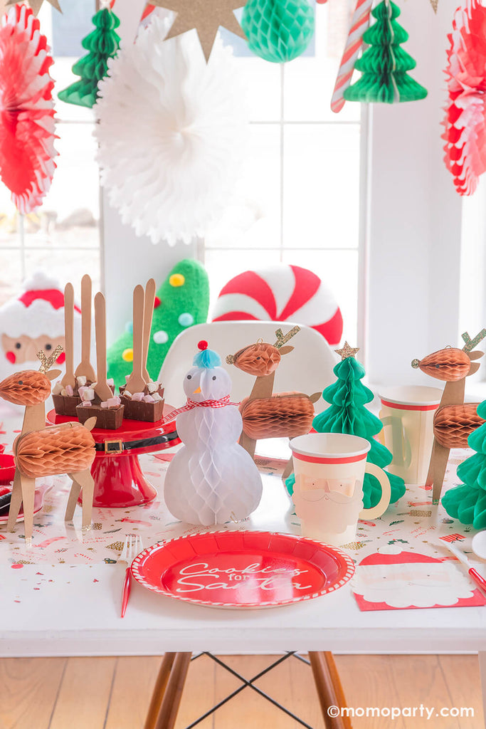 A festive Christmas cookie decorating party table featuring Momo Party's Cookies for Santa red round plates, Santa face party cups, Santa shaped napkins and red and white cutlery set. In the back, there are Christmas character honeycomb featuring Santa, Christmas trees and reindeers. In the back there's a jumbo Christmas honeycomb garland featuring candy canes, peppermints, Christmas trees and shining stars.