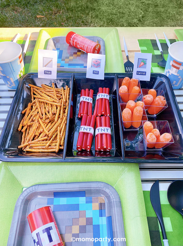 Close up shot of Minecraft themed Birthday Party Sweets and Party Snacks Ideas by Momo Party. A black dividing tray of mini carrots as carrots, pretzel sticks as sticks, and cutted Twizzlers with paper wrap to make TNTs. Surrounding with Minecraft Twist Poppers layered on top of Minecraft themed party plates, pixel TNT theme goodie bags decorated the kids picnic table. 