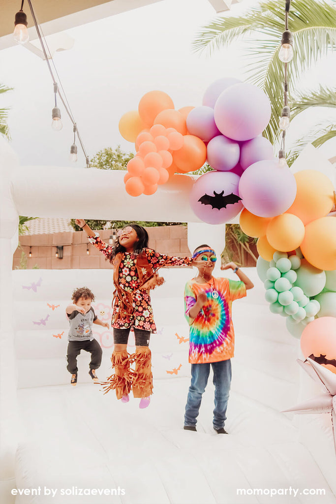 Momo Party Soliza Events Kid's Groovy Halloween Party with Bouncy House and Kid's dressed up in retro hippie outfits and tie-shirt, with a pastel balloon garland decoration on the bouncy house, makes it a perfect inspo for kid's Halloween themed party idea!