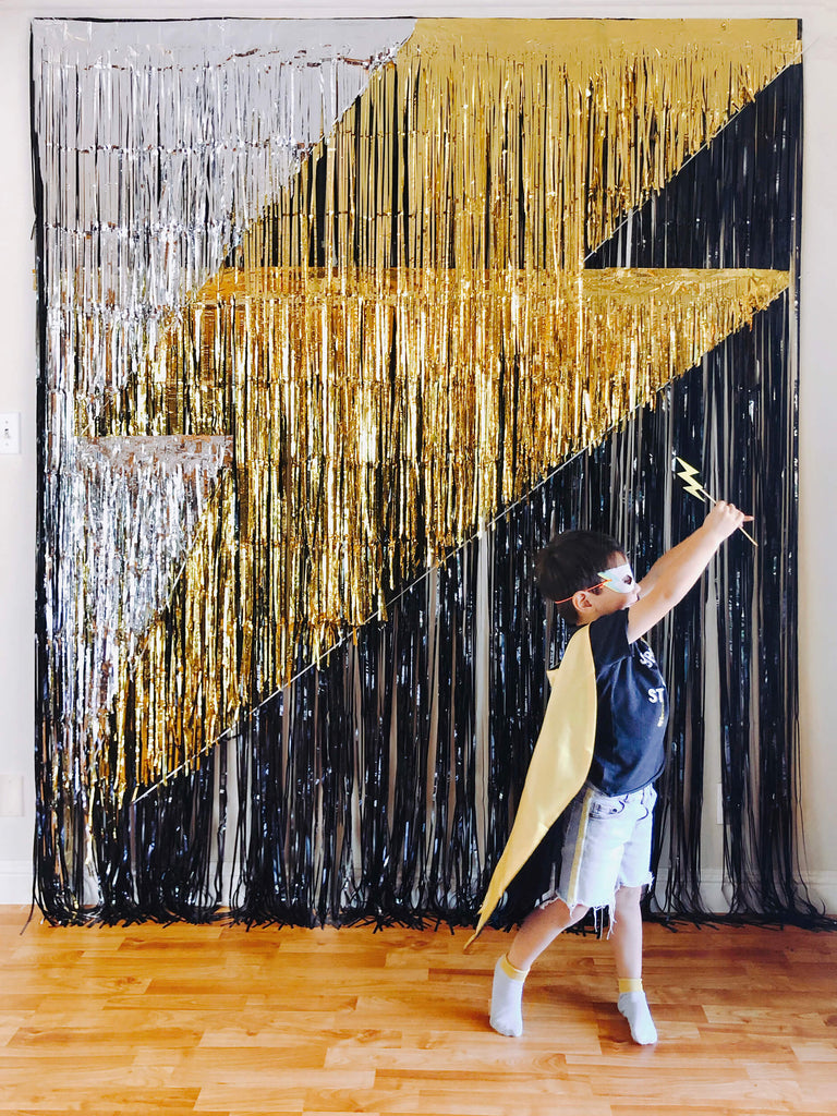 little superhero boy with mask and cape running in front of a black gold sliver fringe curtains made giant lighting bolt icon backdrop