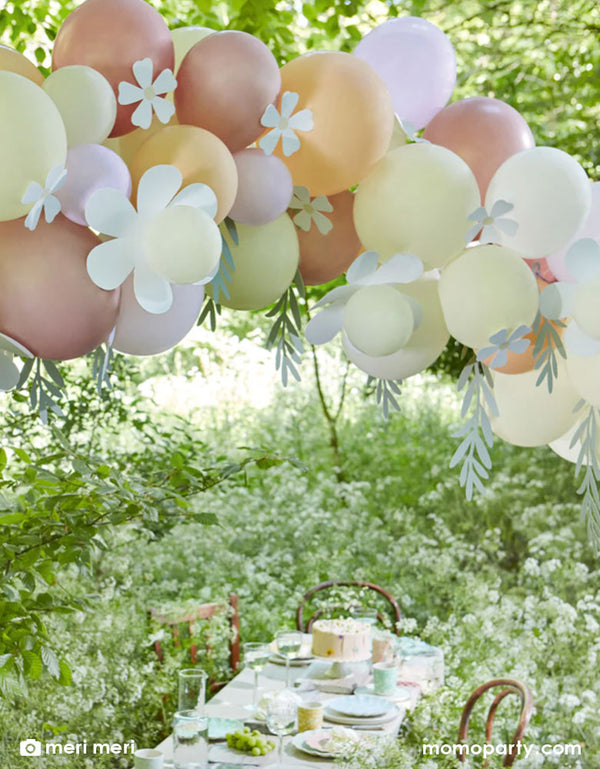 A beautiful party table set up in a field filled with wild daisy flowers. Above the party table it was Momo Party's Pastel Daisy Balloon garland by Meri Meri consisting 51 balloons in white, ivory, lilac, blush and rose gold with daisy shaped paper decorations and foliage shaped paper decorations on it. It makes a perfect decoration for a daisy themed birthday party or a spring inspired celebration.