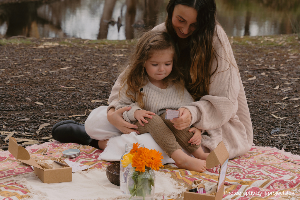 Mother and daughter sitting on a rug reading an affirmation with their potion kit set up all around them. They are out in nature.