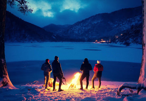 Four men standing around a campfire at night in the winter, with a frozen lake and cottage in the background