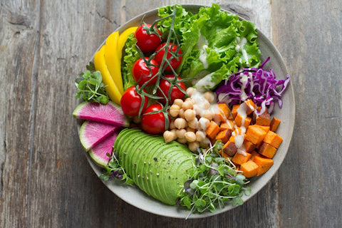 A colorful Buddha bowl filled with assorted vegetables including sliced avocado, red cabbage, chickpeas, cherry tomatoes, yellow bell pepper, and roasted sweet potatoes, arranged on a rustic wooden table.