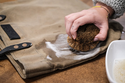 hand holding rough scrub brush, cleaning waxed canvas tote with soapy water