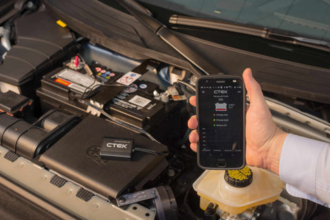 Hand holding a smartphone with the CTEK battery sense app in front of the open hood of a car to check the battery status