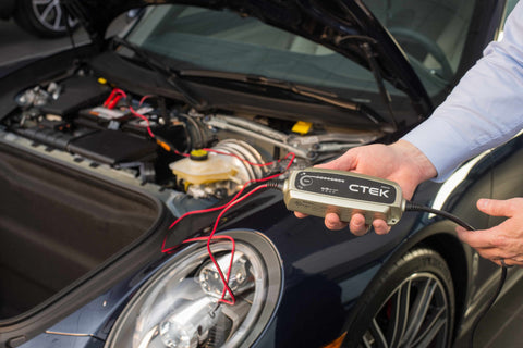 hands holding a CTEK battery charger with an open hood of a car in the background showing the engine