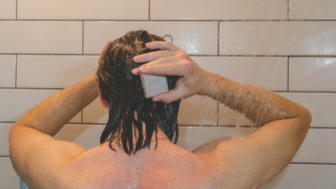 Man washing hair in shower, with white tile. Holding shampoo bar against head.