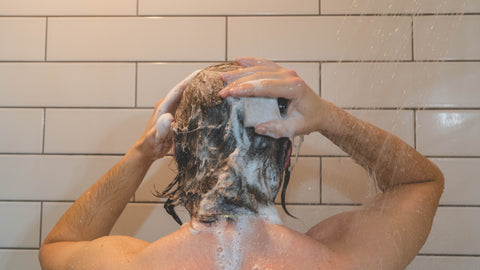 Man washing hair in shower, with white tile. Sudsy hair, holding shampoo bar.
