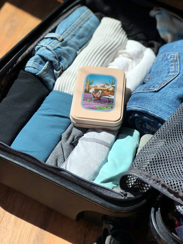 Pink travel tin with lavender, sitting on top of rolled clothes inside of open suitcase.