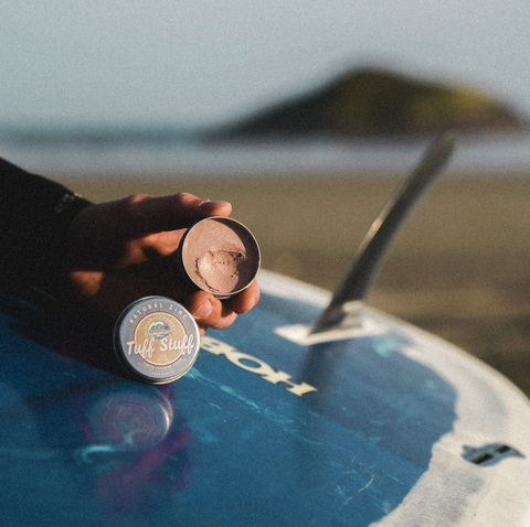 Tin of zinc cream in hands, tin is open, leaning on surf board at the beach