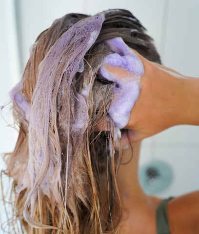 blonde girl washing hair with purple shampoo. Image shows purple suds as girl massages shampoo into scalp.