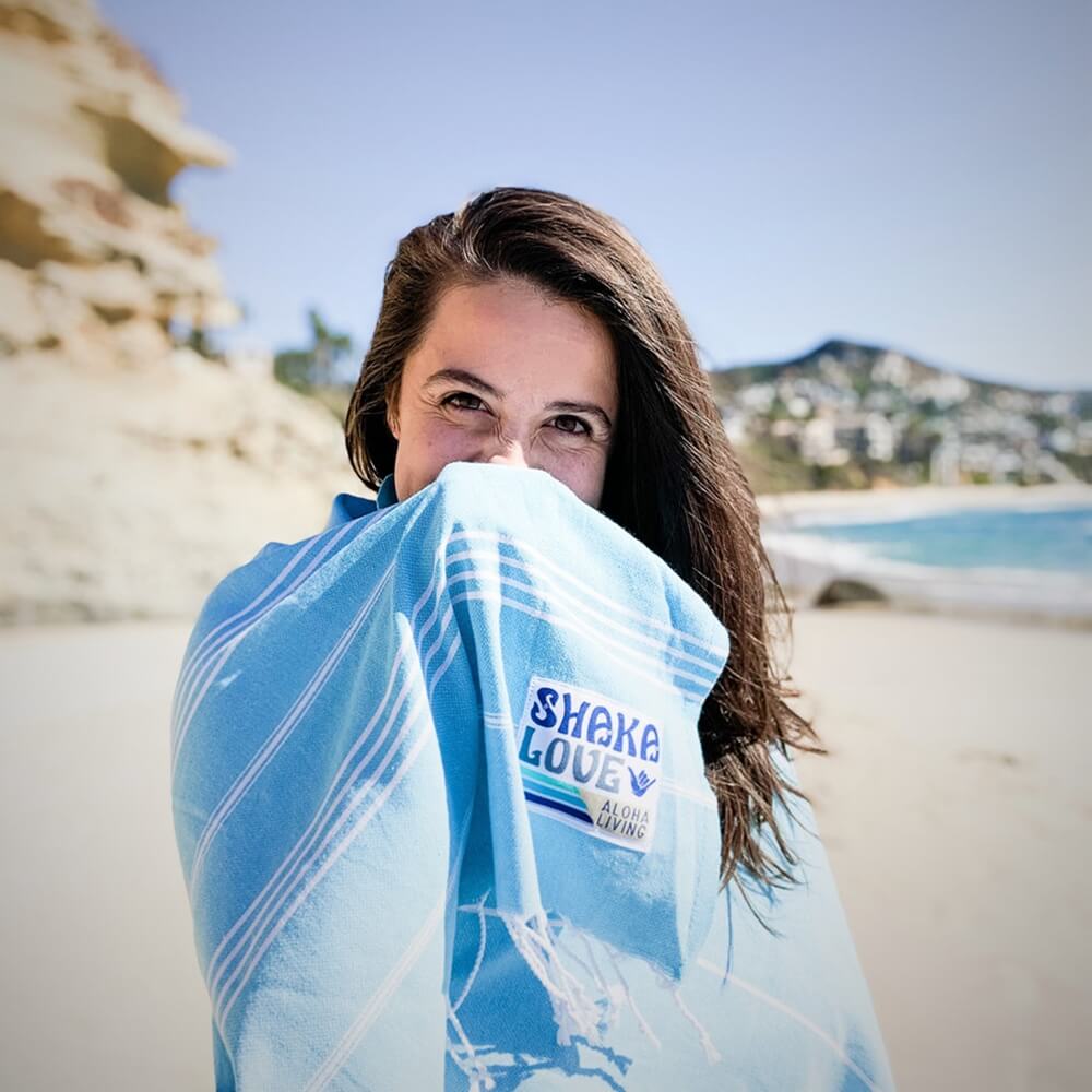 A woman smiles on the sand, spreading out her sustainable Shaka Love beach towel for a day of fun and relaxation