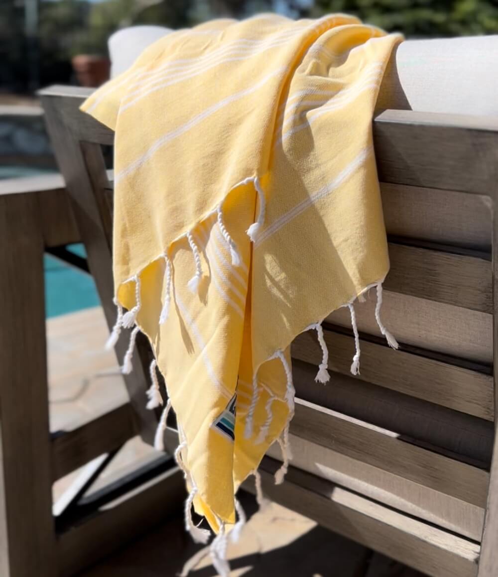 A bright yellow Shaka Love beach towel draped over the back of a wooden chair, next to a sparkling blue pool on a sunny day.