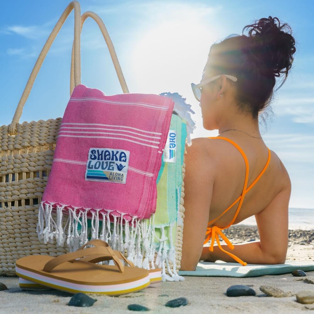 Young woman relaxing at the beach, sitting near a woven bag with two colorful Turkish beach towels resting on top. Her sandals are also visible beside the bag