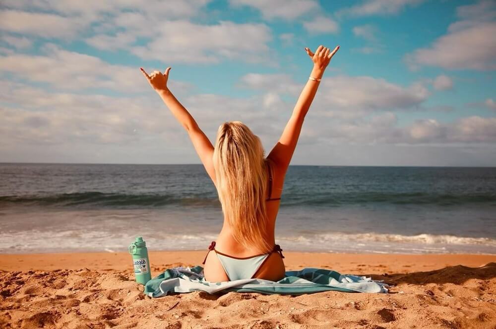 Young woman sitting on a Shaka Love sand-free beach towel, facing the ocean with her arms raised in the air, her joyful expression suggests she's enjoying a carefree day at the beach