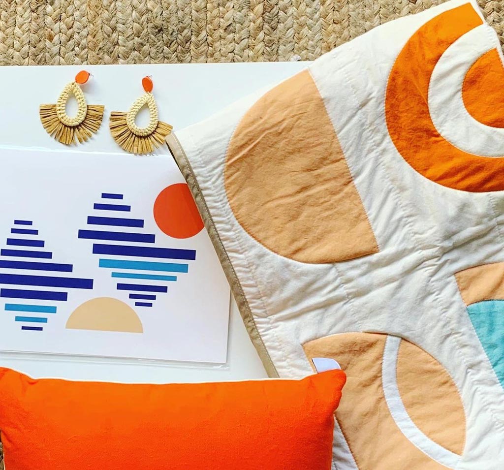 looking down onto a grouping of objects: a quilt with orange designs on it, and orange cushion, a little piece of artwork with oranges and blues