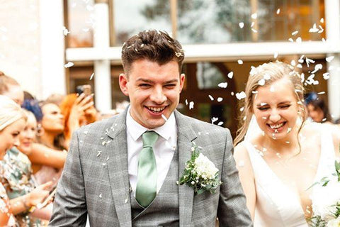 A groom wearing a sage green tie, smiling joyfully as confetti is thrown around him, adding a vibrant and celebratory atmosphere to the wedding scene.