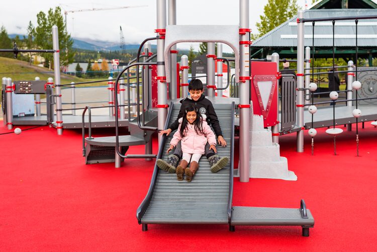 Two kids slide down the Roller Slide with Transfer Bench at the Jumpstart Inclusive Playground in Whitehorse, YT.