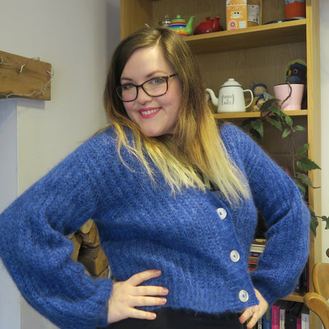 Hannah, a mid-size female with long brown hair fading to blonde ends, wears a fluffy blue cardigan and poses with her hands on her hips.