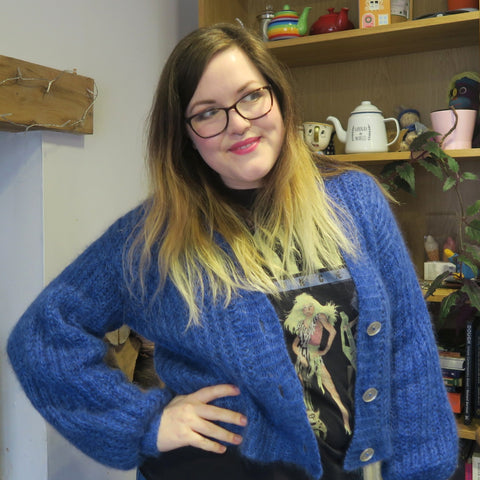 Hannah, a mid-size lady with long hair going from brown roots to blonde ends, wearing a vibrant blue, fluffy cardigan unbuttoned, with a hand on her hip.