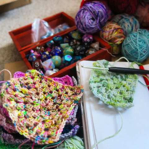 A box of dice sits behind a stack of colourful, crocheted granny squares.