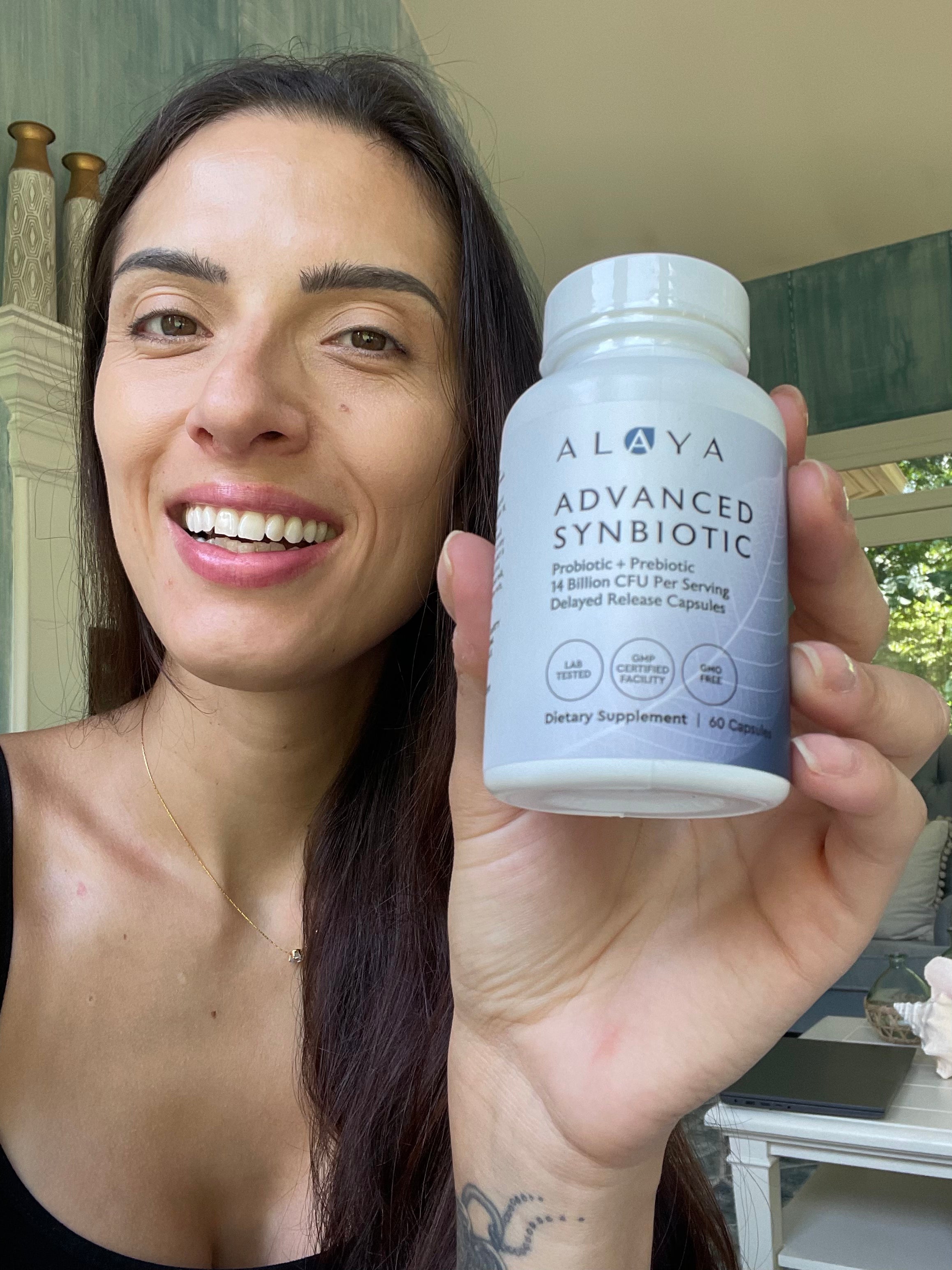 A woman holding a bottle of Alaya Advanced Synbiotic dietary supplement and smiling.