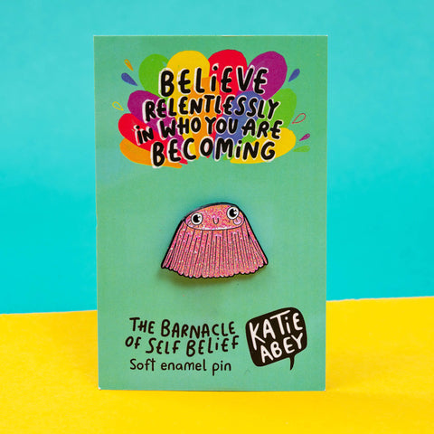 The Barnacle of Self Belief Enamel Pin Badge on its green backing card with a rainbow splash at the top with black text reading 'believe relentlessly in who you are becoming' and bottom black text reading 'the barnacle of self belief, soft enamel pin' and the Katie Abey Logo, on a yellow and teal blue background. A pink glittering smiling barnacle pin with big eyes, eyelashes and pink cheeks all with a black outline. Hand drawn design by Katie Abey.