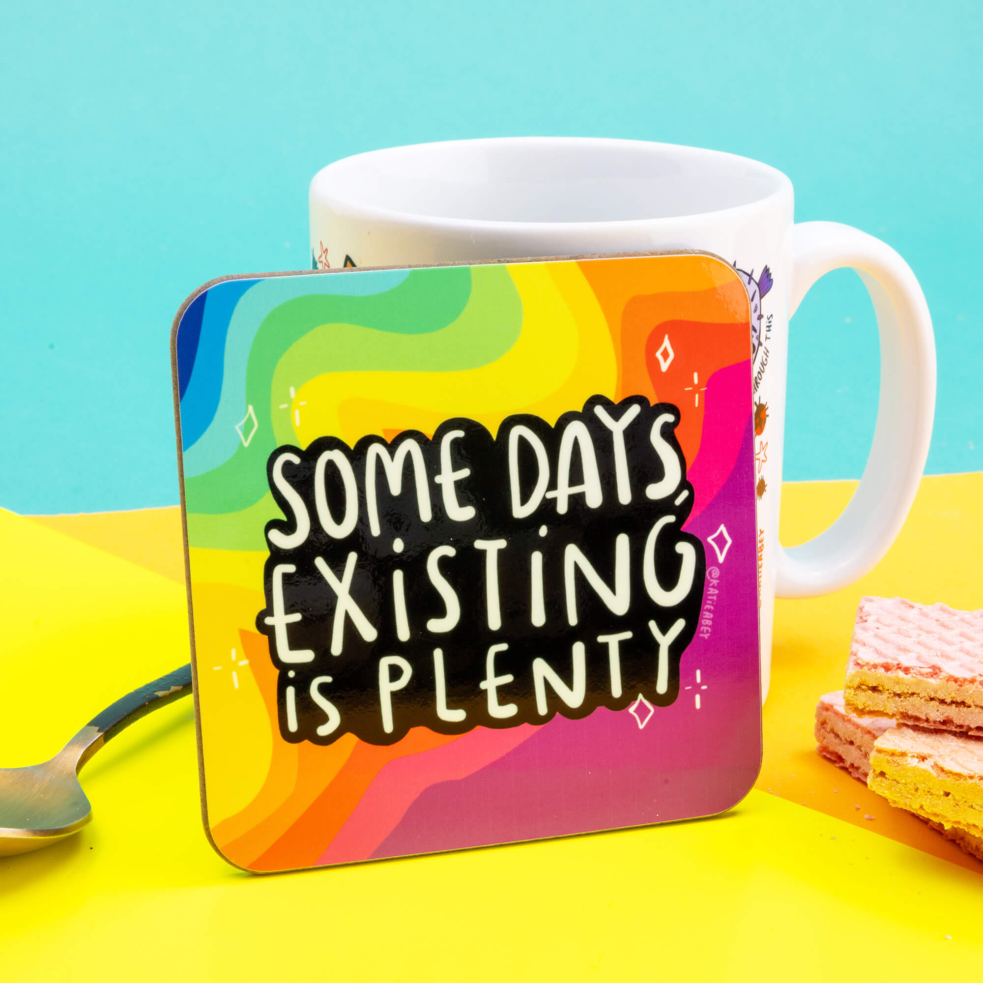 A coaster designed and illustrated by Katie Abey with rainbow background and text saying, 'some days, existing is plenty'