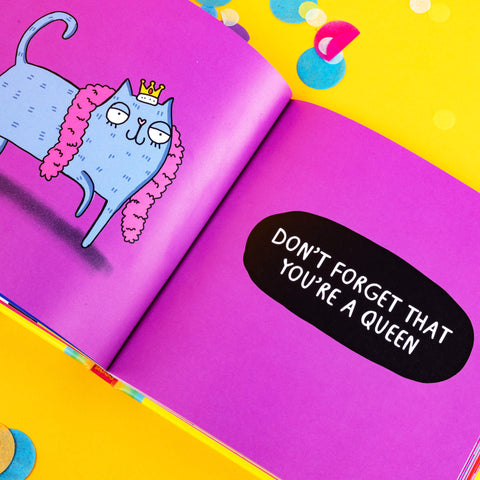 an illustration of a grey cat with a smug look and is wearing a crown and pink feather boa. The other page has text that reads DONT FORGET THAT YOURE A QUEEN. The book is laid on a bright yellow background amongst coloured confetti.