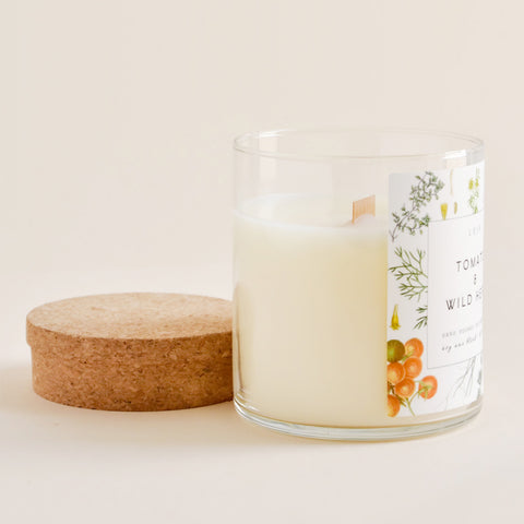 A glass candle with botanical label and wooden wick sits next to its cork lid.