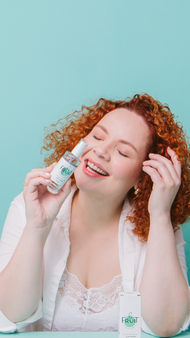 Mulher branca, ruiva, vestindo um robe branco, sorri de olhos fechados enquanto segura um desodorante de uva verde em uma das mãos