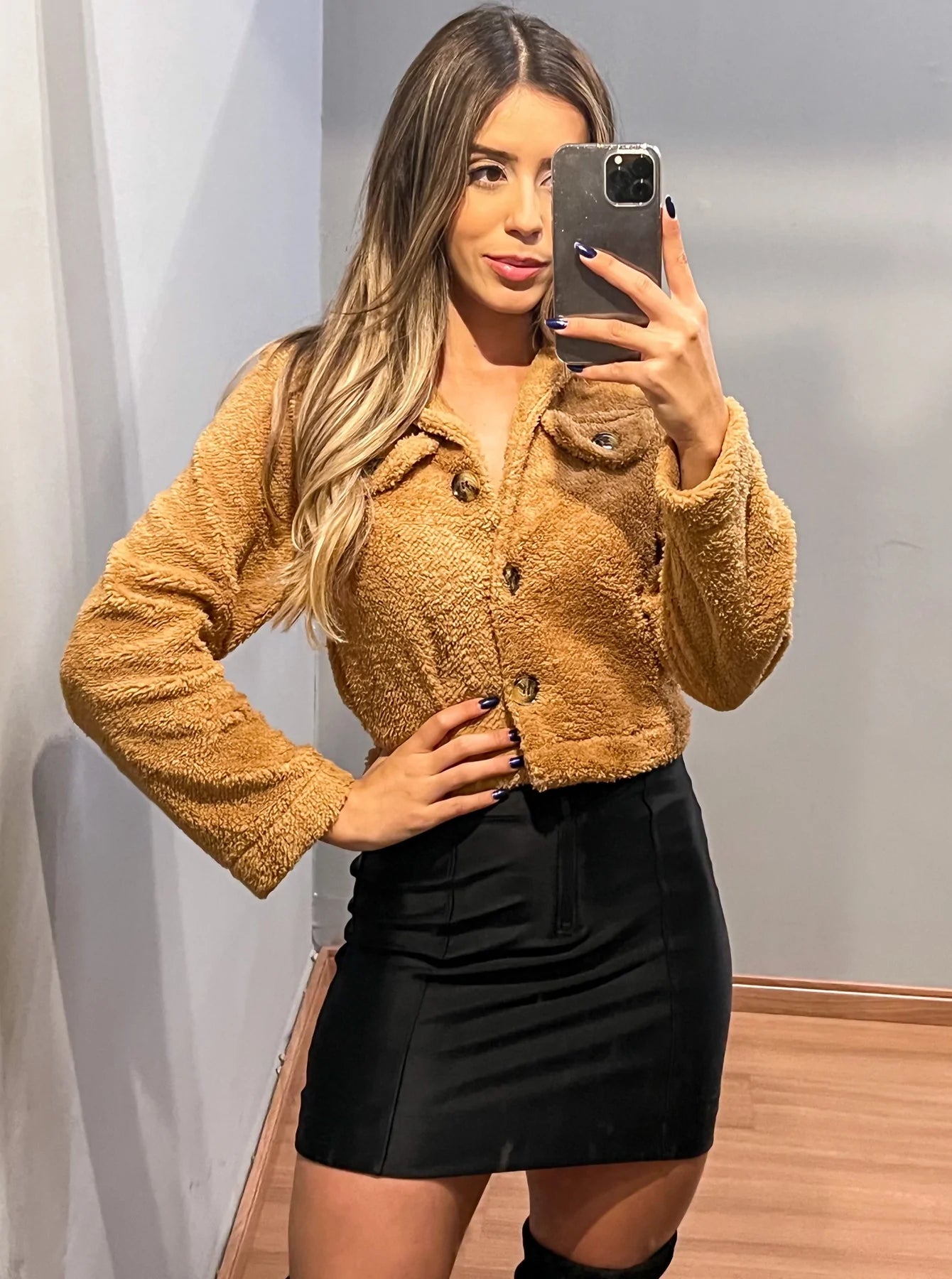 Selfie de modelo em frente a um espelho posando com um casaquinho teddy marrom, saia preta e botas de cano alto pretas.